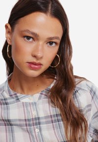 Geruite shirt in lichte kleuren met patronen in blauw, roze en wit. Ronde halslijn, knopen aan de voorkant en een zachte textuur. Model draagt creolen.