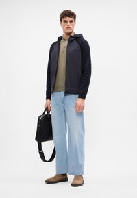 Casaco com capuz preto e azul-marinho com mangas texturizadas, camisa polo verde-oliva, calças de ganga largas azuis claras e botas de camurça castanhas; a carregar uma bolsa preta.