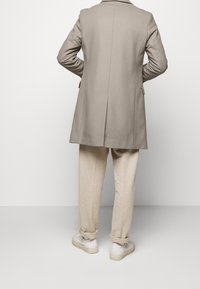 Cappotto beige su misura con uno spacco sul retro, abbinato a pantaloni di lino beige chiaro e sneakers bianche, visto da dietro.