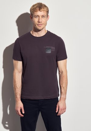 T-shirt à manches courtes de couleur violet foncé avec un col rond et un détail d'étiquette imprimée sur la poitrine. Le modèle se tient devant un fond uni.