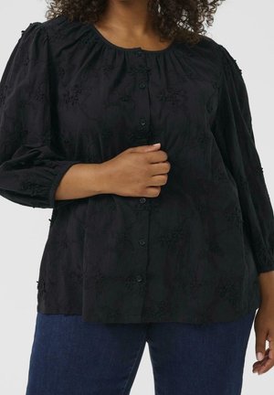 Kvinde iført en sort bluse med lange ærmer med blomsterprint og blå jeans, hånd hviler på blusens knapper.