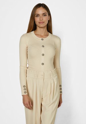 Frau trägt eine beige gerippte, geknöpfte Strickjacke und hochtaillierte, plissierte cremefarbene Hosen, steht vor einem schlichten weißen Hintergrund.