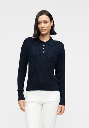 Granatowy dzianinowy polo z teksturowanym wzorem, czterema złotymi guzikami i prążkowanymi mankietami. W połączeniu z białymi spodniami, posiada małe logo na piersi.