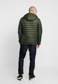 Jachetă puffer de culoare verde măsliniu cu glugă ajustabilă, textură matlasată și fermoar. Asortată cu blugi închiși la culoare și adidași negri.