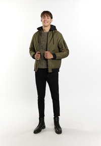 Giacca bomber verde oliva con cappuccio nero, chiusura con zip e tasche laterali, abbinata a un maglione di maglia e pantaloni neri; stivaletti neri con lacci.