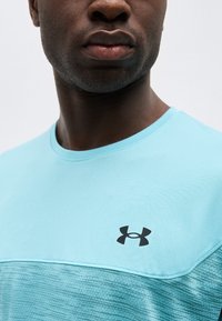 Κοντινό πλάνο ενός άνδρα που φοράει γαλάζιο αθλητικό πουκάμισο με υφή και μαύρο λογότυπο Under Armour στο στήθος.