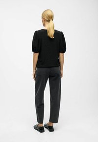 Kvinde med blondt hår i hestehale iført en sort top med pufærmer, mørke lige ben jeans og sorte loafers, står med ryggen til.