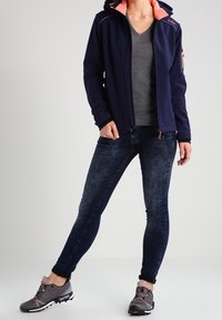 Dunkelblaue Softshelljacke mit einer pinken gefütterten Kapuze, kombiniert mit einem grauen V-Ausschnitt-Shirt, dunklen Jeans und grauen Sportschuhen mit weißen Sohlen.