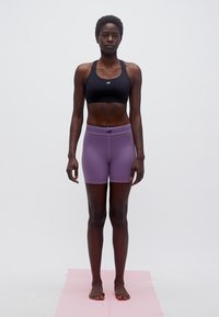 Γυναίκα που στέκεται ξυπόλυτη σε ροζ χαλάκι γιόγκα, φοράει μαύρο αθλητικό σουτιέν και λιλά σορτς γυμναστικής, με φόντο απλό λευκό.