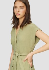 Jasnozielona sukienka kopertowa wykonana z teksturowanej tkaniny, z krótkimi rękawami, przewiązywanym w pasie oraz guzikami wzdłuż przodu.