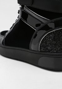 Gros plan d'une basket montante noire avec cuir verni brillant, panneau de talon pailleté et semelle en caoutchouc épaisse sur fond blanc.