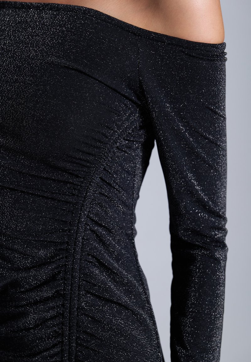 Robe noire décolettée à épaule dénudée avec un design froncé, fabriquée dans un tissu texturé et brillant, présentant de longues manches et une silhouette ajustée.