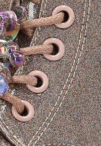 Zapato de tela brillante marrón con ojales rosa claro. Presenta cordones trenzados y acentos de cuentas coloridas. Vista en primer plano de los detalles del diseño.