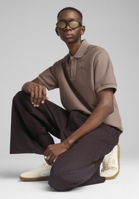 Lys brun polo skjorte med tekstureret stof og korte ærmer, parret med mørke bukser og off-white sneakers. Har tonede solbriller.