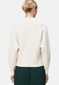 Pull crème à côtes avec un design court, des manches longues et un tissu texturé. Porté sur une jupe plissée verte. Finition simple et épurée.