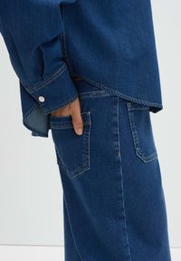 Donzen overhemd in donkerblauw met lange mouwen, puntige kraag en metalen knopen; gecombineerd met donkerblauwe denim broek met achterzakken.