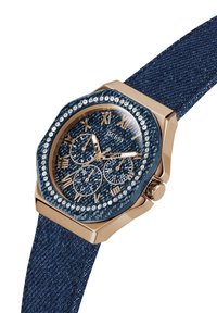 Oktagonal klocka med en denimrem och en blå texturerad bezel, med guldtonade romerska siffror och en ring av pärlor runt kanten.