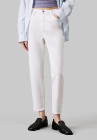 Jeans in denim bianco con gamba dritta, vita alta e cinque tasche, abbinati a una camicia a righe blu chiaro e scarpe slip-on nere.