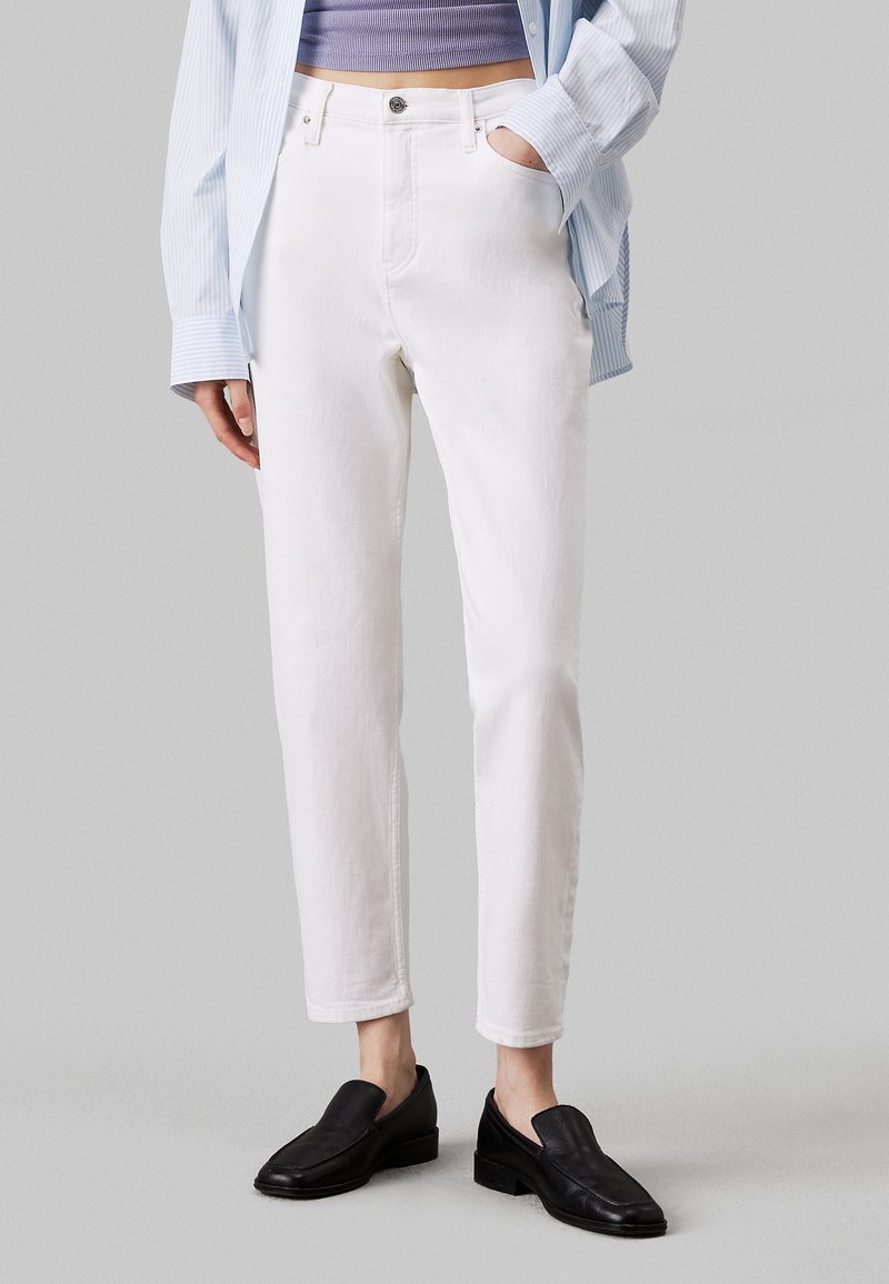Jeans in denim bianco con gamba dritta, vita alta e cinque tasche, abbinati a una camicia a righe blu chiaro e scarpe slip-on nere.