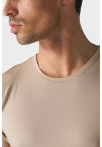 Beżowa dopasowana koszulka z okrągłym dekoltem. Gładka tekstura materiału z subtelnymi detalami szycia wzdłuż kołnierza. Zbliżenie na ramiona.