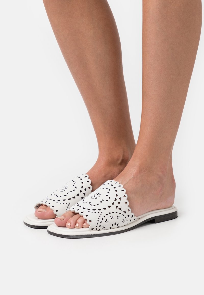 Jambes portant des sandales blanches à enfiler avec des bords festonnés et des motifs floraux perforés complexes, debout sur une surface blanche.