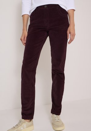Femme portant un pantalon en velours côtelé violet foncé avec des poches devant et des baskets beiges, debout sur un fond clair uni.