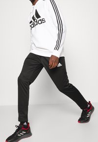 Černý Adidas teplákový set s bílým logem, skládající se z bílého mikinového svršku s kapucí a černých zúžených kalhot. Černé sportovní boty s červenými akcenty.
