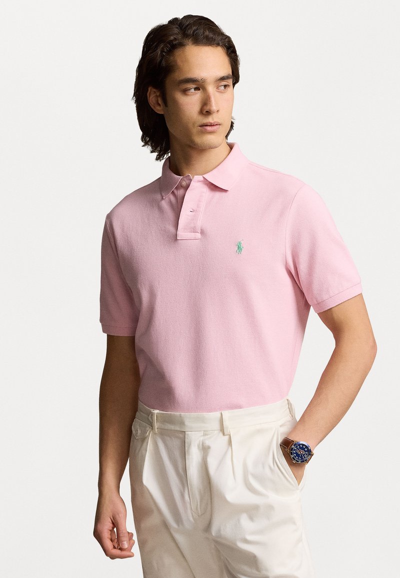 Tricou polo roz deschis, dintr-un material texturat, cu mâneci scurte și un placket cu nasturi din lemn; prezintă un logo brodat verde pe piept.
