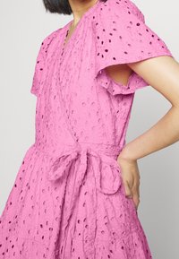 Růžové šaty z texturované tkaniny s detaily ve formě očíček, krátkými volánkovými rukávy, zavinovacím designem a uvázaným pasem.