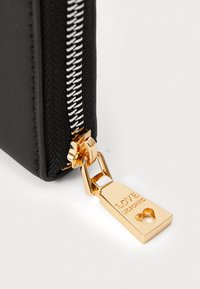 Carteira preta com fecho de correr, exterior texturizado, apresentando um puxador de metal dourado e nome da marca gravado. Close-up do detalhe do fecho.