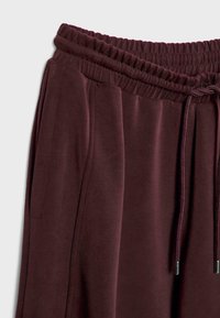 Maroon trenirke s elastičnim pojasom, zatvaranjem s špagom i bočnim džepovima. Glatka tekstura, suženi dizajn i minimalni detalji šivanja.