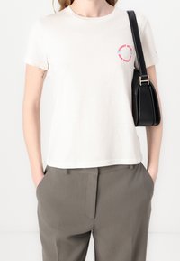 White cotton t-shirt with short sleeves and a round neckline. Features red and blue text logo on the front. Paired with grey trousers.