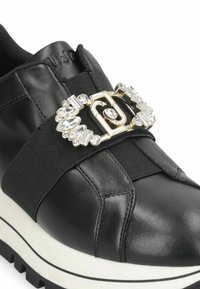 Baskets en cuir noir avec une bande élastique et une boucle métallique décorative ornée de gemmes transparentes. Semelle texturée blanche avec des accents noirs.