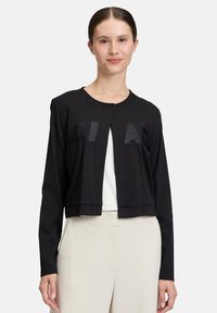 Kobieta w czarnej, długiej kardiganie na guziki z małymi ćwiekami i tłoczonymi literami, w połączeniu z jasnobeżowymi spodniami o wysokim stanie.