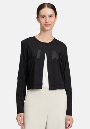 Donna che indossa un cardigan corto nero a maniche lunghe con piccoli borchie e lettere in rilievo, abbinato a pantaloni beige chiaro a vita alta.