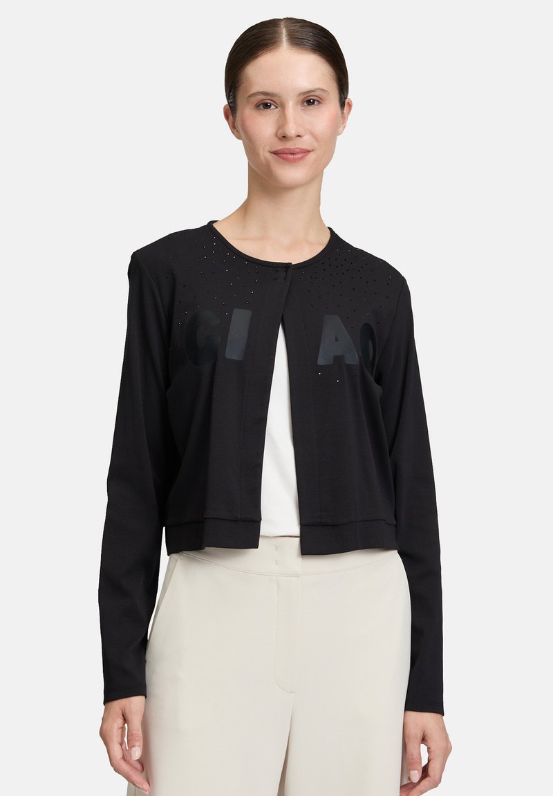 Kobieta w czarnej, długiej kardiganie na guziki z małymi ćwiekami i tłoczonymi literami, w połączeniu z jasnobeżowymi spodniami o wysokim stanie.