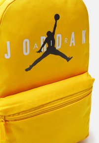 Zaino di tessuto giallo con logo Jumpman nero e testo "JORDAN" bianco. Include una tasca frontale con zip e finitura testurizzata.