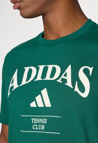 T-shirt de algodão verde com o ousado texto branco "ADIDAS" e um gráfico de um logótipo triangular, com "TENNIS CLUB" impresso abaixo em uma caixa retangular.