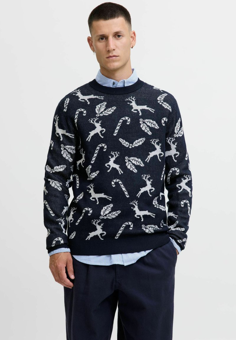 Pull en tricot bleu marine avec des motifs de rennes blancs, de cannes en sucre et de feuilles. Encolure ronde et poignets côtelés, associé à une chemise collée bleu clair.