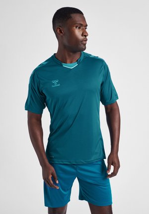 Mann in türkisfarbener Sportbekleidung, schaut nach rechts und steht mit entspannter Haltung vor einem schlichten hellen Hintergrund.