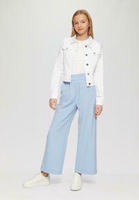 Weiße Jeansjacke über einem weißen Top, kombiniert mit hellblauen, weiten Hosen. Das Model trägt weiße Sneakers; der Hintergrund ist neutral.