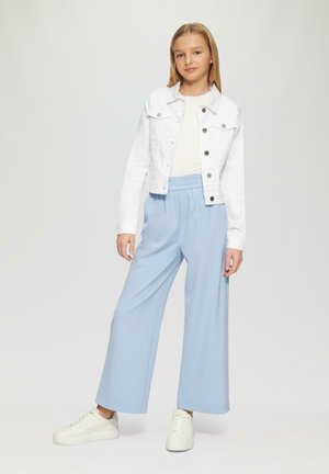 Witte denimjas over een wit topje, gecombineerd met lichtblauwe wijd uitlopende broek. Model draagt witte sneakers; achtergrond is neutraal.