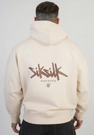 Person mit tätowiertem Hals und Händen trägt einen hellbeigen Hoodie mit dem Schriftzug „SikSilk Ride or Die“ auf dem Rücken, von der Kamera weggewandt.