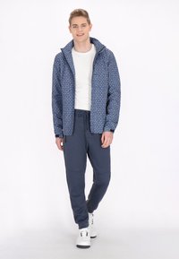 Ținută masculin cu o jachetă albastră cu model și pantaloni jogger bleumarin. Jacheta are glugă și fermoar în față. Pantalonii jogger sunt conici și au buzunare.
