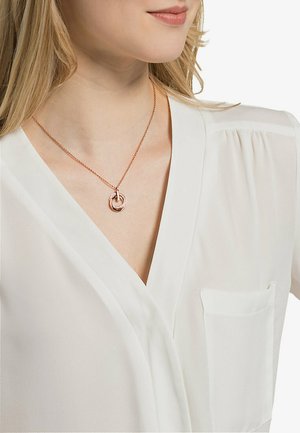 Collana in oro rosa con anelli intrecciati, caratterizzata da una superficie texture, sospesa a una fine catena, indossata su una camicetta bianca.