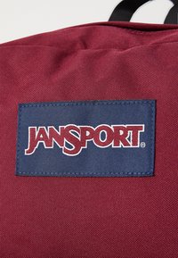 Sac à dos en tissu rouge avec un patch rectangulaire bleu portant l'inscription "JANSPORT" en lettres blanches. Surface texturée avec des coutures visibles.