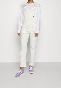 Salopette di denim bianco con spalline regolabili, tasca frontale e accessori in metallo argento, abbinata a una camicia bianca a maniche lunghe e sneakers viola.
