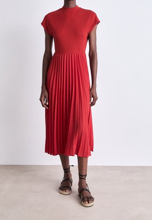 Red pleated midi dress with a high neck and short sleeves. Smooth, textured top merging into flowing pleats. Worn with black strappy sandals.