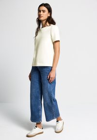 Haut crème à manches courtes associé à un jean en denim bleu à jambes larges et des baskets blanches. Le haut a un col rond simple et une texture lisse.