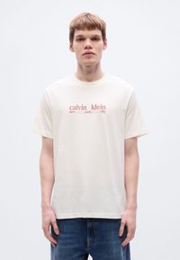 Μπεζ βαμβακερό μπλουζάκι με κοντά μανίκια, με κόκκινη έντυπη γραφική επιγραφή: "calvin klein new york city" στο κέντρο του στήθους.